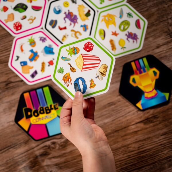 Player holding a hexagonal Dobble Connect card with colorful symbols like popcorn, dice, and guitar. Other Dobble Connect cards are laid out on a wooden table.